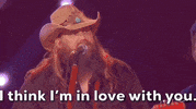 ACM Awards gif. Man in tan cowboy hat with guitar strapped around him sings "I think I'm in love with you" into mic stand.