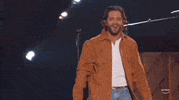 Academy of Country Music Awards gif. Male singer in suede button-up walks past on stage pianoist and in front of guitarist as he lists arm to bring microphone closer to his mouth which carries a smiling expression.