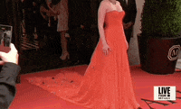 Met Gala 2024 gif. Nell Diamond poses on the red carpet at the entrance to The Mark wearing a cherry red Hill House Home sleeveless gown with a structured bodice fitted through the waist and a full flowing skirt and with flower detailing at the end of the long train. 
