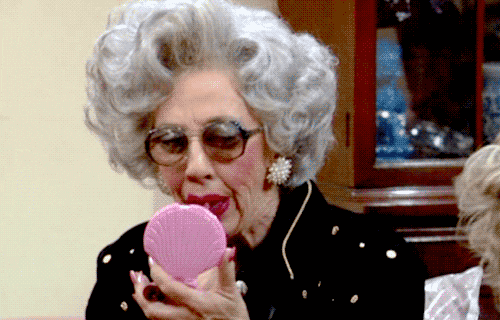 Video gif. An elderly woman with a huge mop of curly gray hair delicately applies lipstick to her already-red lips with a mirrored compact. 