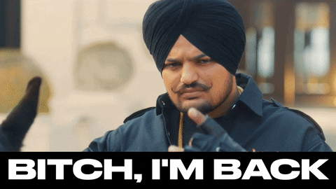 Celebrity gif. Rapper Sidhu Moosewala gazes outward and flashes a signature hand gesture with thumb and middle finger folded down. Text, "bitch, I'm back."