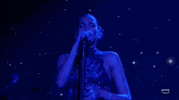 ACM Awards gif. While gripping mic stand with one hand Dua Lipa waves free hand at ear-level height as she sings and lightly shakes her head from side-to-side. Her eyes are closed as if it helps her sings. 