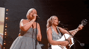 TV gif. The musical duo Tigirlily Gold, wearing gray dresses, singing and playing guitar enthusiastically on stage at the 2024 ACM Award show.