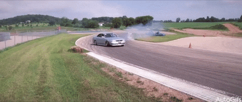 Video gif. A silver lowrider sedan drifts around a curb, kicking up a cloud of dust.