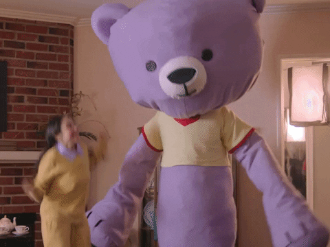 Video gif. A gigantic lavender teddy bear in a pale yellow heart t-shirt sways cheerfully in a living room. A girl in a yellow outfit next to him jumps up and down excitedly, and the room seems to be shaking from their movement. Text, "Congrats!"