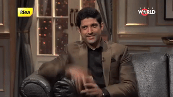 TV gif. Farhan Akhtar, on Koffee with Karan, sits on a couch and raises his pointer finger to the air, smiling and glancing off to the side.