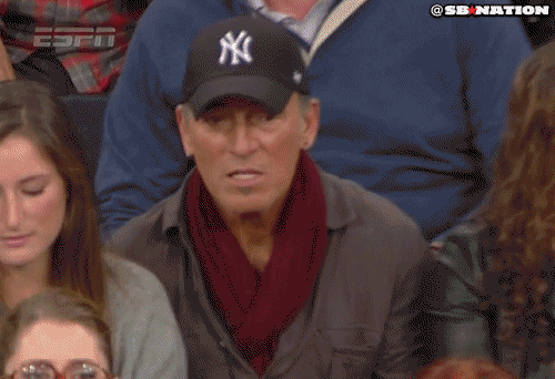 Sports gif. Bruce Springsteen sits in the stands at a Duke-UCLA game and wears a New York Yankees baseball cap. He pumps his fist as a celebratory gesture.