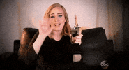 Celebrity gif. Sitting on a sofa, Adele holds up a trophy in her hand and waves hello with her other hand, smiling.