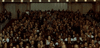 Video gif. In an ornate performance hall a well dressed crowd stands with a standing ovation. 