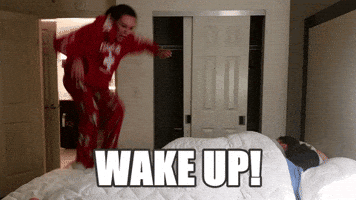 Video gif. Woman in a red tracksuit jumps up and down on a bed and wakes up a couple sleeping under the covers. Text, "Wake up!"