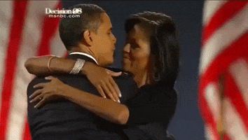 Political gif. Barack and Michelle Obama stand in front of American flags. Michelle's arms are wrapped around his shoulders as they kiss, then she puts her face to his cheek and pats his shoulder. 