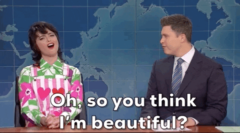 SNL gif. Melissa Villasenor and Colin Jost on the Weekend Update sit next to each other. Melissa looks up, batting her eyes, and holding her hands together firmly on the desk. She says, “Oh, so you think I'm beautiful?” Colin Jost laughs. 