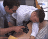 Video gif. Two men are in an office and one is asleep at his desk. The other staples the sleeping man's tie to the desk then slaps him awake. The man jolts awake and tries to attack his friend but gets caught by the staple and falls down.