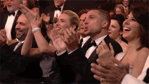 TV gif. A camera zooms out on a large crowd of celebrities sitting at an award show. The front row claps, sitting down, and laughing with wide smiles on their faces. The rows behind them stand up to give a standing ovation, cheering on the person on stage with immense enthusiasm.