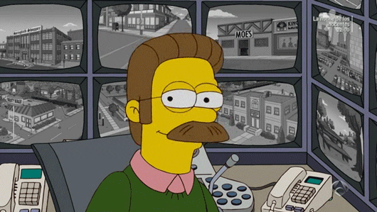 Simpsons gif. Ned Flanders sits at a desk and looks at us before giving us a wide, closed mouth smile.