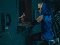 Music video gif from Sabrina Carpenter's music video for "Please Please Please." She wears a blue dress with a hood around her head, dark sunglasses, and elbow length black gloves. She is visiting Barry Keoghan in jail. He is behind glass in an orange jumpsuit. They both pick up a telephone and talk.