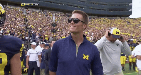 Sports gif. Tom Brady waves from the field of the University of Michigan football stadium as the crowd applauds.