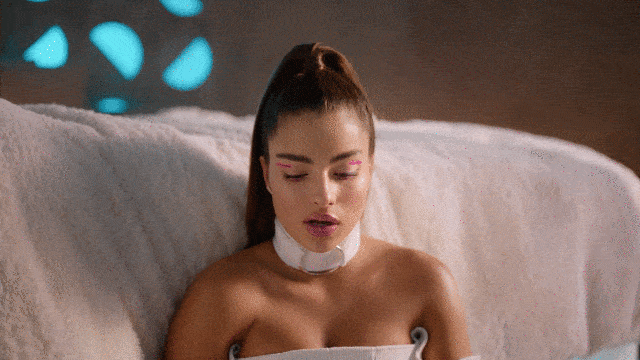 Music video gif. From video for Thought About That, Noa Kirel slowly lifts her eyes open to look at us while speaking, sitting on a plush white couch.