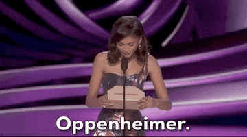 Oscars 2024 gif. Zendaya wears a glimmering, purple holographic dress that coordinates with the royal purple backdrop. She reads off of the envelope and announces Oppenheimer for Cinematography. 