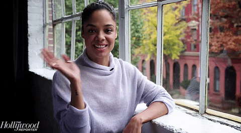 Celebrity gif. Leaning against a window sill, Tessa Thompson smiles and waves at us.