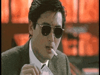 Video gif. A man in a suit wearing sunglasses lights a cigarette in his mouth with a $100 bill that is on fire.