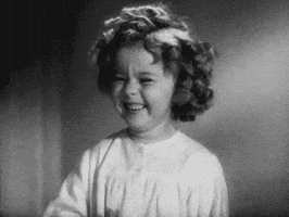Celebrity gif. Black and white shot of Shirley Temple scrunching her nose and having a big laugh, covering her mouth with her hand.