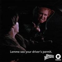 Movie gif. Bette Midler as Winifred and Omri Katz as Max in Hocus Pocus. She flies on a broom as she keeps up with a car on the road and peers into the driver's seat where Max is driving. He looks terrified as he looks at her and back on the road and she says, "Lemme see your driver's permit!"