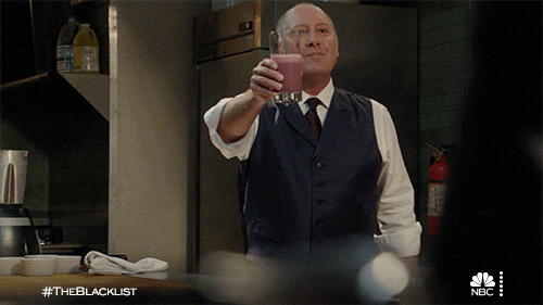 TV gif. James Spader as Red in The Blacklist. He is wearing a suit in a kitchen and he smiles and lifts a glass filled with a smoothie in cheers before bringing it to his lips to take a sip.