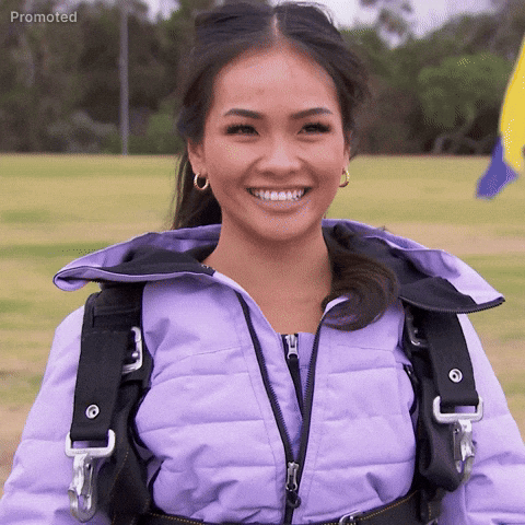 Sponsored gif. Jenn Tran, the bachelorette from season 21 of The Bachelorette, has a full teeth smiling smirk on her face, sticking her tongue out mischievously, eyes darting playfully from side to side. Tran stands on a field of grass in front of a yellow and blue flag wearing a harness over her coat.