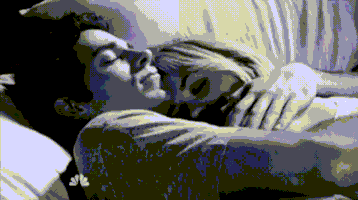 TV gif. A black-and-white scene of a couple cuddling in bed. A man lying on his back has his arms wrapped around a woman, and he turns slightly to kiss her forehead.