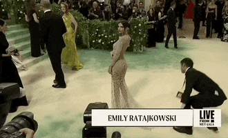 Met Gala 2024 gif. Emily Ratajkowski wearing a sandy sheer Versace gown, strikes a bold, wide-legged over-the-shoulder pose.