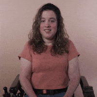 Reaction gif. A Disabled white-presenting Latina woman with cerebral palsy and kinky-curly brown hair, seated in her motorized wheelchair, hugs herself, smiles, and leans in with an impish sweetness.