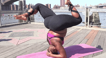 Sports gif. Jessamyn Stanley, a yoga instructor with a curvy body type, does a headstand while touching her feet together slowly on a yoga mat on a rooftop, showing her strength and control. 