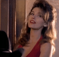 Celebrity gif. Shania Twain wearing a red dress and red lipstick and leans against a wall while looking at someone sexily before tilting her head down and saying, "Babyyyyy."