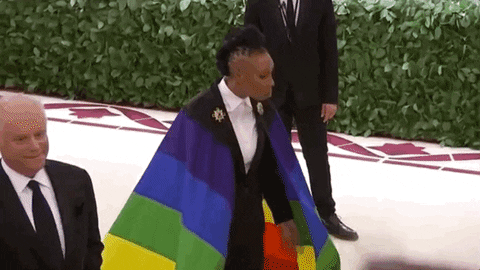 Celebrity gif. Lena Waithe wears a rainbow cape pinned to the shoulders of her suit on the Met Gala red carpet while lights flash from everywhere.
