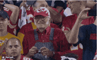 Video gif. Standing in the audience of a football game, an old man wearing overalls and a Kansas City Chiefs hat bites his bottom lip as he squints out at the field. He then holds up a small digital camera up to his face and squints into it to take a picture.