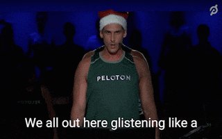 Ad gif. A Peloton ad of a muscly man in a Peloton tank top with a headset microphone and a Santa hat on says, “We all out here glistening like a beautiful Christmas ham right now.”