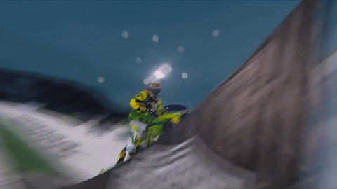 Sports gif. At the X Games, a snowmobile driver goes up a ramp, and as the vehicle does a 360-degree flip he turns around on it so that he is facing backwards and raising his hand when it lands on the snow.