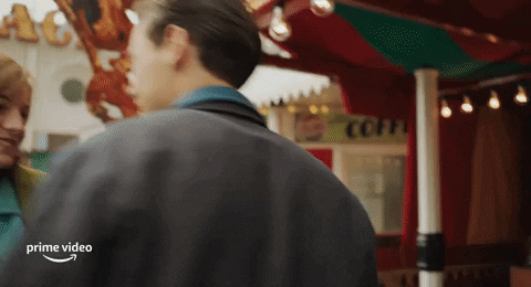 Movie gif. Harry Styles as Tom in "My Policeman," stands outside of a carnival game cheering and celebrating with his arms open wide, then kisses the girl next to him on the cheek.