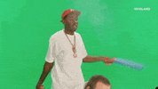 Celebrity gif. Tyler the Creator stands in front of a greenscreen and swings a flimsy toy sword to fan away smoke, which makes text reading, "Welcome Back" appear.