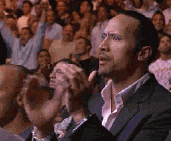 Celebrity gif. Dwayne “the Rock” Johnson stands up in a crowd of people chewing on gum and slowly clapping. 