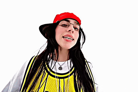 Music Video gif. From Billie Eilish's music video for "LUNCH", Billie is on a stark white background dressed in a white long sleeve shirt and a yellow and black jersey over the top, a red flat brim hat on. She is walking backwards while staring into the camera and singing.