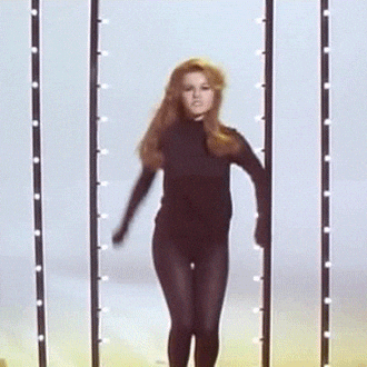 Video gif. A woman in all black dances, bringing her arms up and over her head to the beat. 