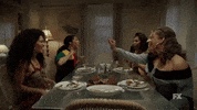 TV gif. The girls from Pose are sitting at a dining table and they all chat enthusiastically while laughing and high fiving one another.
