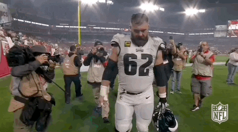 Sports gif. Jason Kelce of the Eagles walks off the field and looks up while raising his helmet to acknowledge the crowd and camera. 