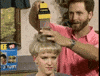 Slow motion infomercial features a man in a pink collared shirt looking mesmerized as he uses a handheld Flowbee hair vacuum to cut and suck the hair of a woman with a sort of bowl cut hairstyle. The woman gives a sharp side eyed expression like she’s checking to see if anyone’s laughing at her. 