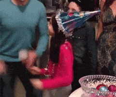 Video gif. Young girl in a pink turtleneck and a sparkling birthday cone hat is overcome with excitement at what appears to be a family birthday. A man yells something towards us behind her and, all of a sudden, she picks up a bowl of colorful balls and smashes it on the floor in front of her, raising her arms in the air like she's totally pumped about it.