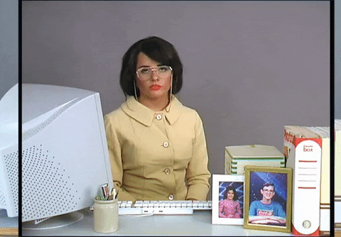 Video gif. A bored-looking receptionist sitting at a desk with a large 90s computer monitor, keyboard, large books and picture frames of children in front of 90s style graphics.