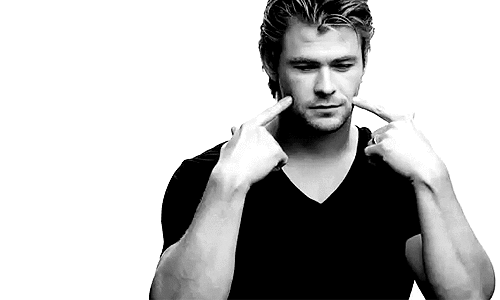 Video gif. A black-and-white clip of Chris Hemsworth putting his fingers to his cheeks as he smiles.