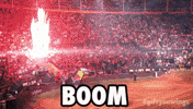 Sports gif. At the edge of dirt bike arena, near a crowded audience, a fiery explosion bursts upward as a motocross driver exits a ramp and soars high above the arena.